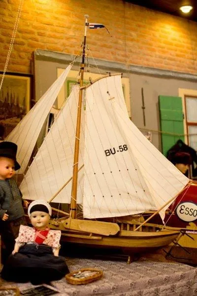 Miniatuurbotter met wit zeil in het museum, met een kinderpop ervoor.
