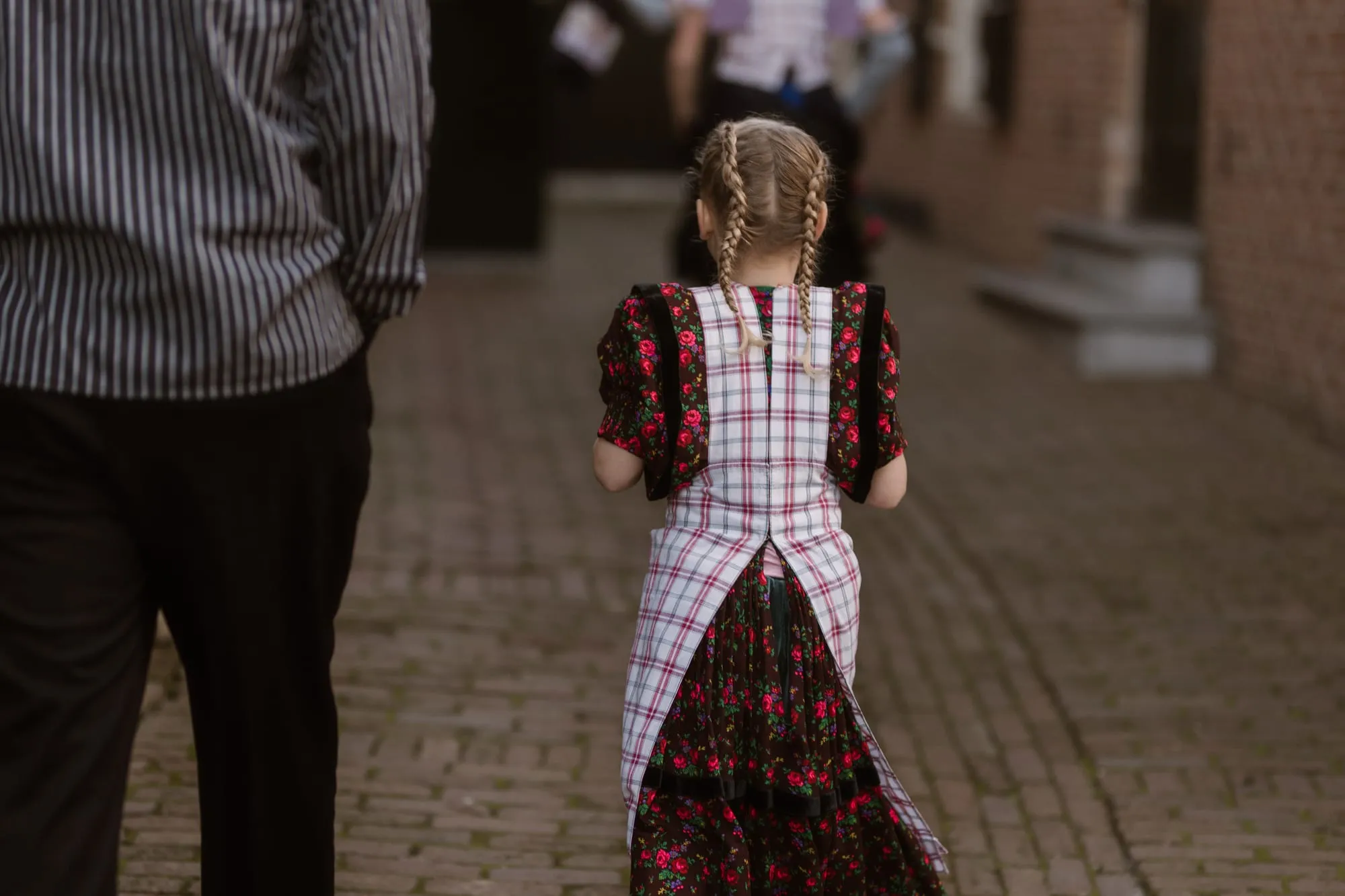 Een meisje met vlechten in kinderdracht met geruit schort loopt over de klinkers, naast een man in gestreept hemd.