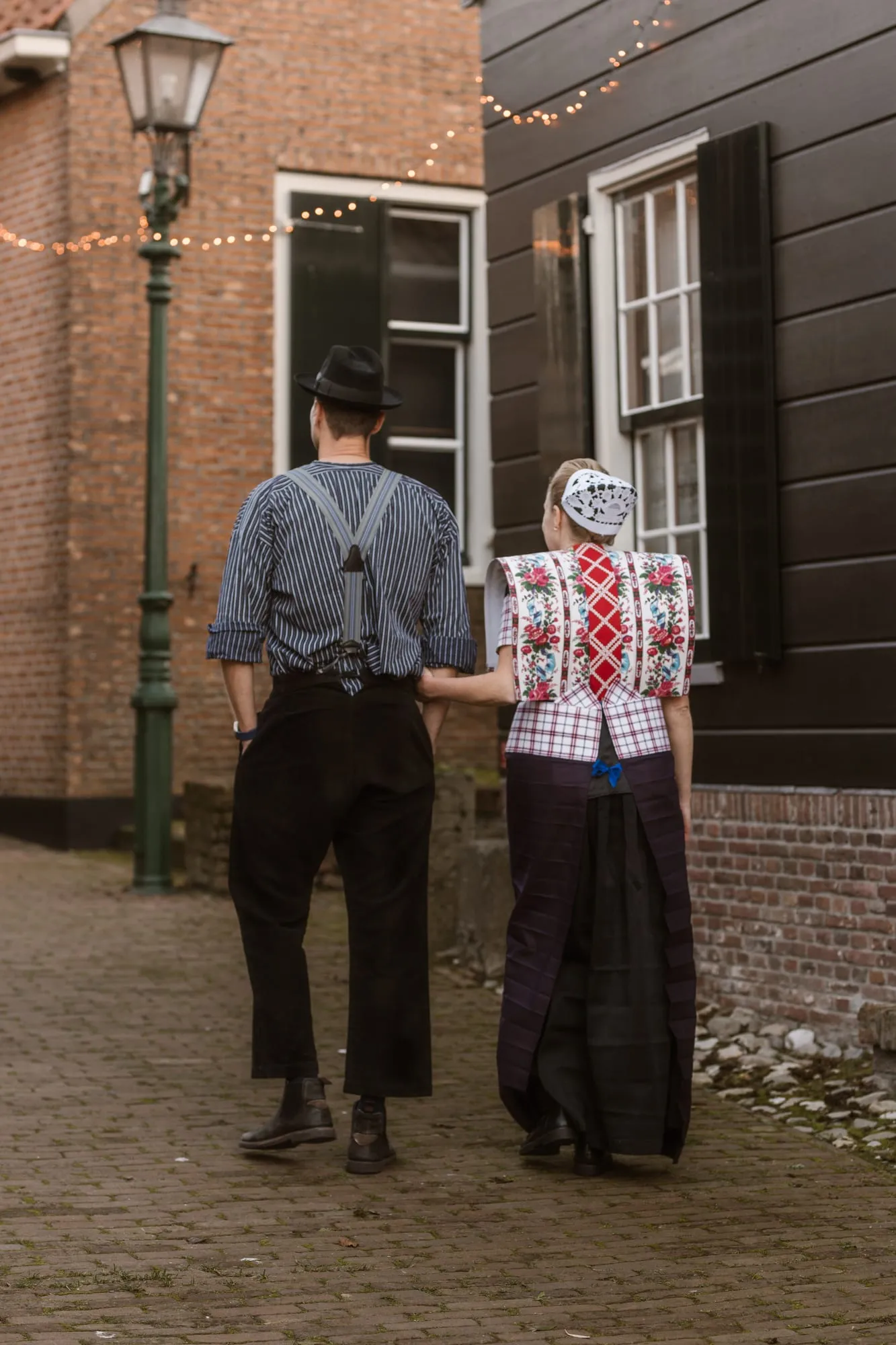 Een man in gestreept hemd met bretels en een vrouw in dracht lopen door een steegje met een lantaarn en lichtjes.