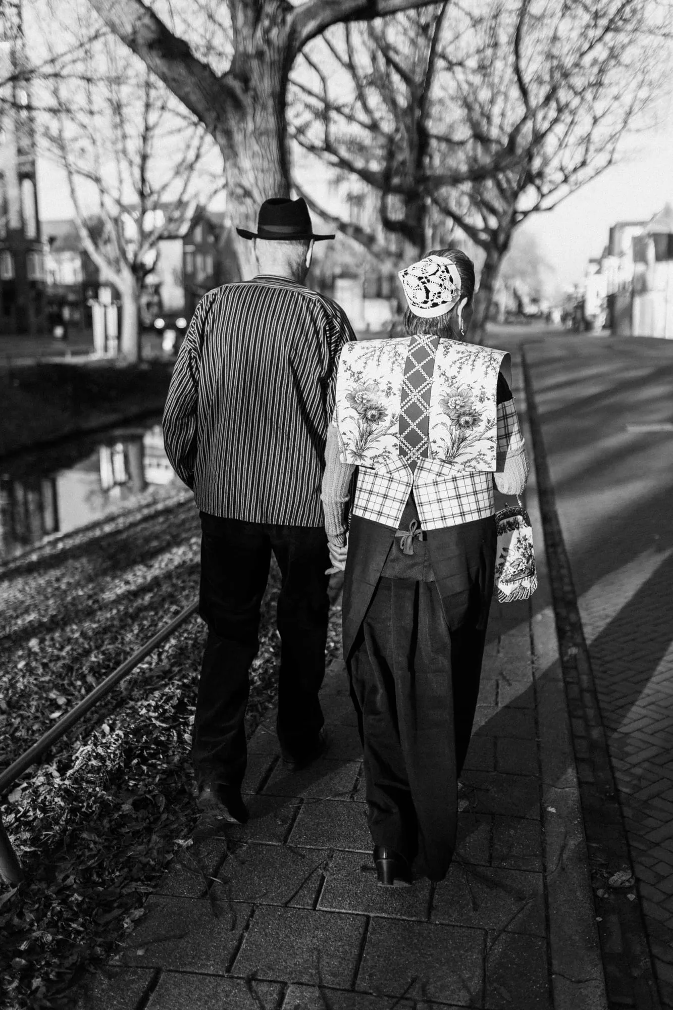 Zwart-witfoto van een man met hoed en een vrouw met kraplap die samen langs de gracht wandelen.