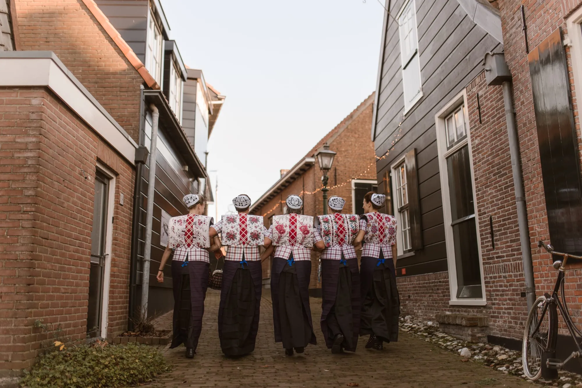 Vijf vrouwen in Spakenburgse dracht lopen gearmd door een steeg tussen bakstenen en houten huizen.
