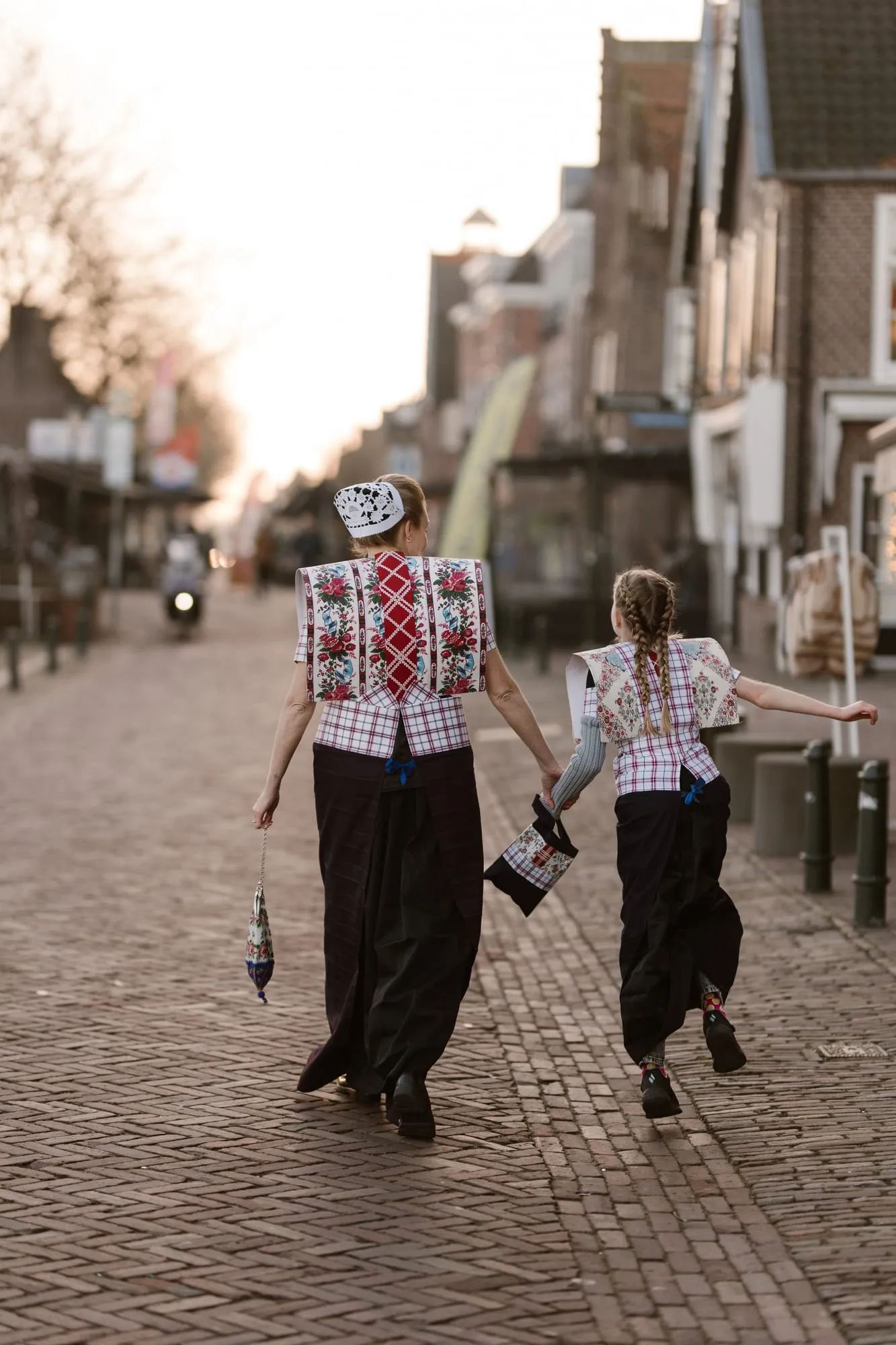Een vrouw en een meisje in Spakenburgse dracht lopen hand in hand door de winkelstraat, op de rug gezien.