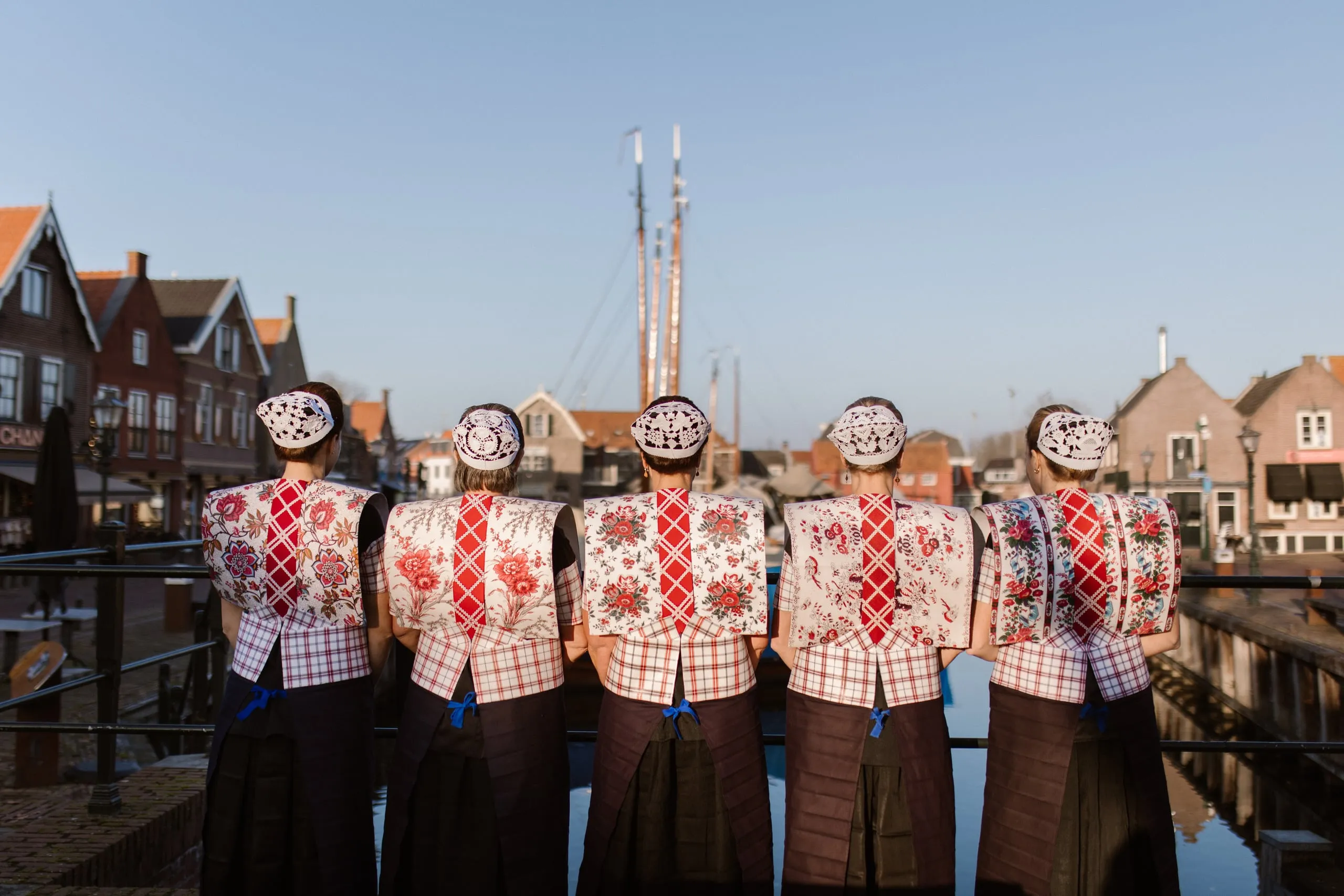 Vijf vrouwen in Spakenburgse klederdracht staan met de rug naar de camera aan de haven; op elke kraplap loopt de rode geruite baan midden over de rug.